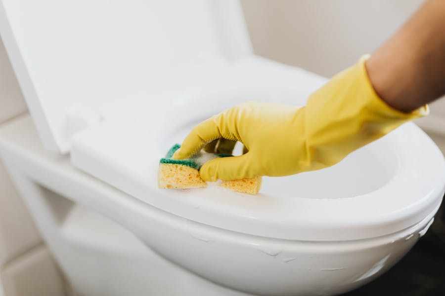 Yellow-gloved hand scrubbing the rim of a white toilet with a sponge for deep clean your house room by room
