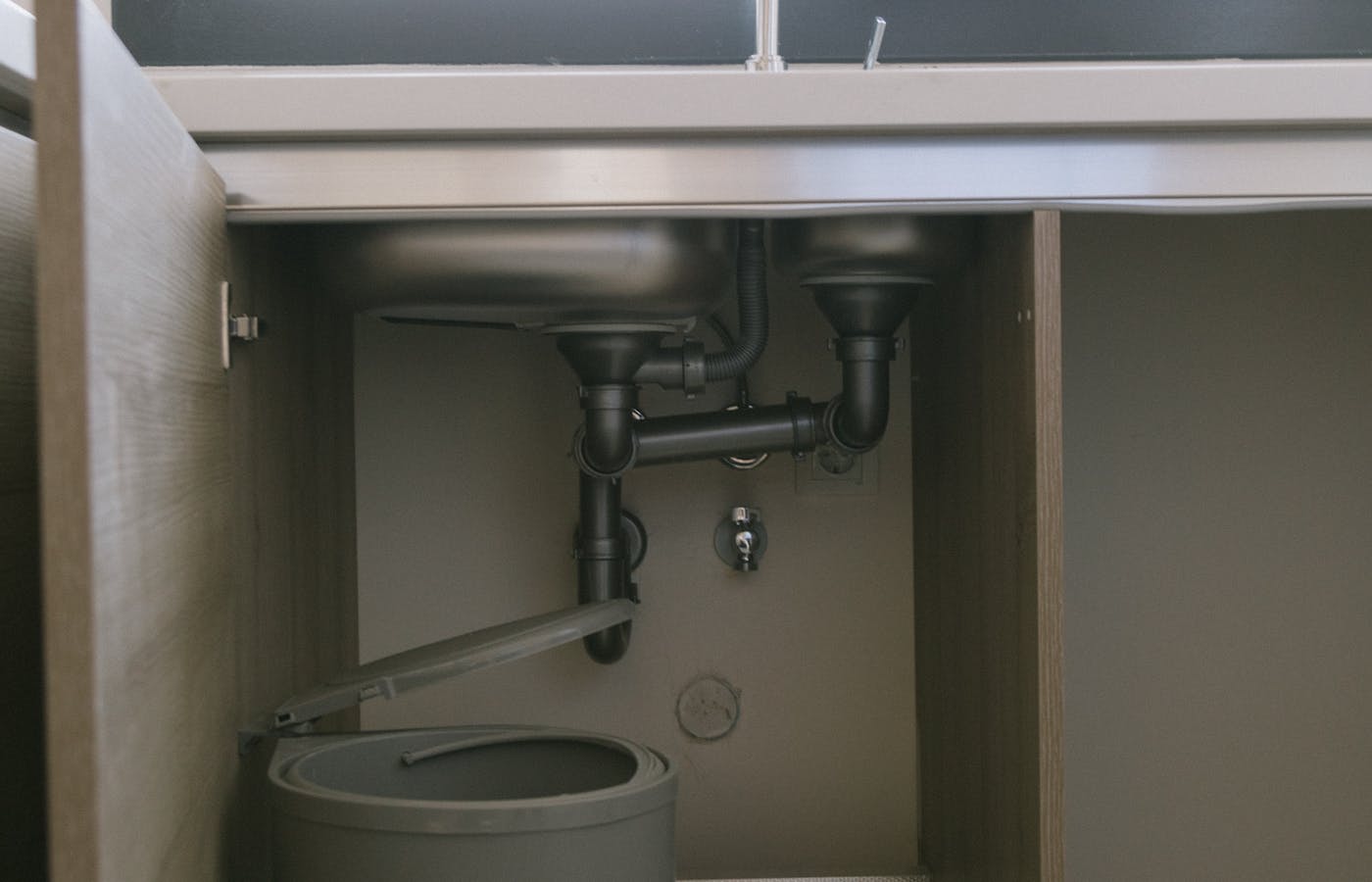 Open under-sink cabinet with visible pipes and a trash bin