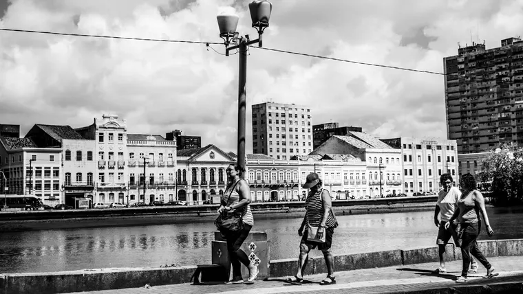 Pessoas caminhando em um parque urbano com área verde e água ao fundo